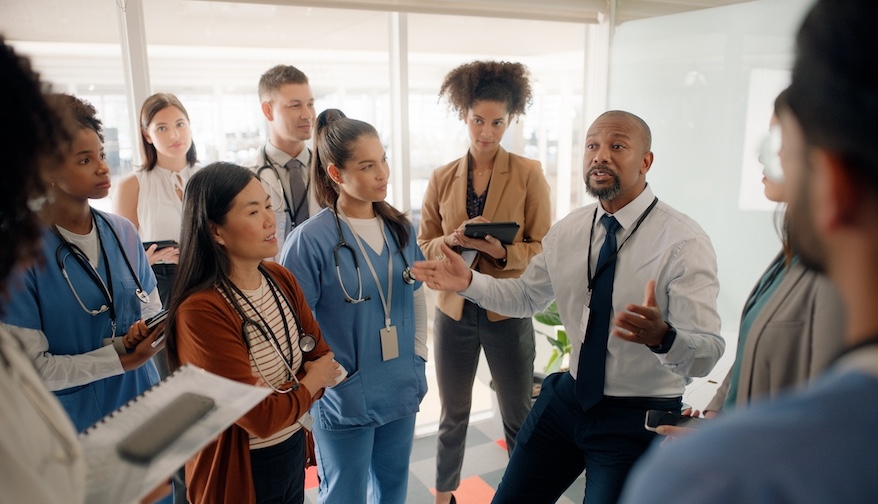 Boardroom, discussion and motivation for staff by executive in hospital for healthcare. Man, training and mentor of employees or nurses and tablets for notes of speech by leader in medical field
